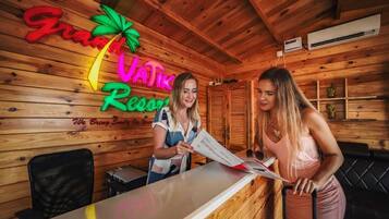 A wooden interior with a reception desk, a neon sign reading "Grand Vatik Resort," and a black leather couch.