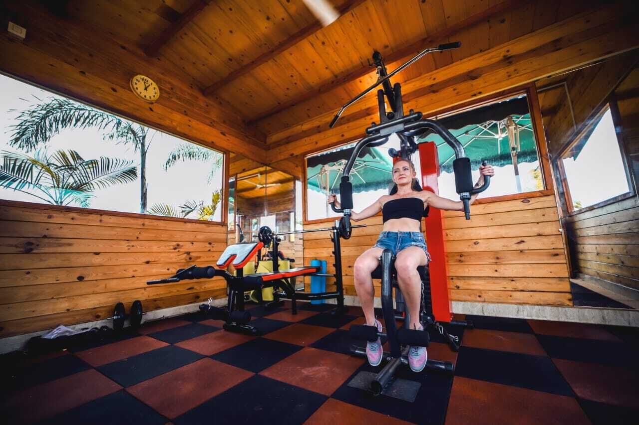 Un gimnasio con paredes de madera y suelo a cuadros, con equipo de ejercicio y una ventana que ofrece vistas a palmeras.