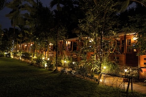 A row of wooden buildings with warm lighting, surrounded by lush greenery and a pathway. - Grand Vatika Resort (Arambol)
