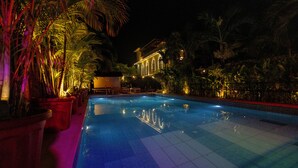 Ein Swimmingpool mit gefliestem Boden, umgeben von Kübelpflanzen und Palmen, mit einem Gebäude im Hintergrund.