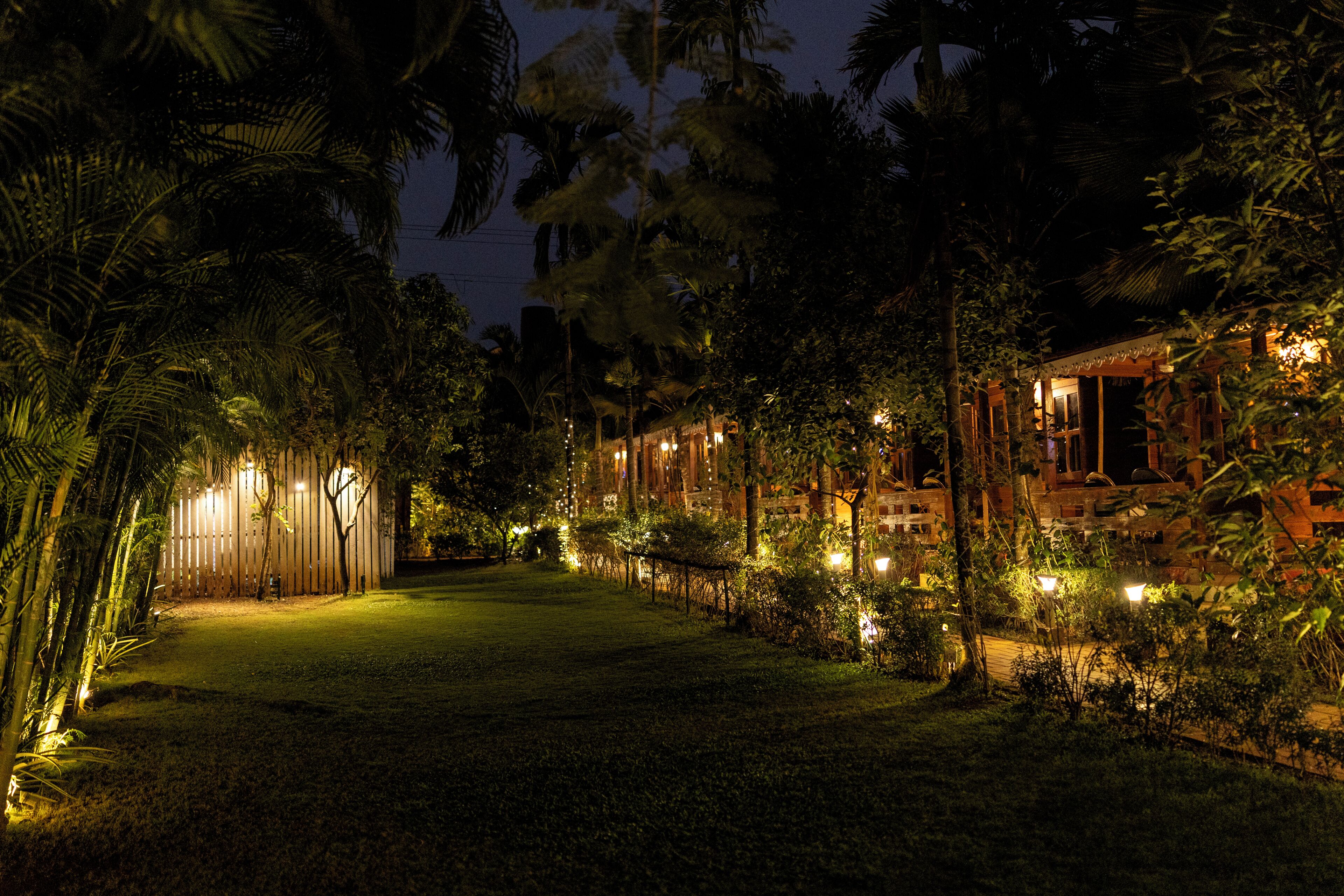 Une vue nocturne d’un hôtel, avec un chemin bien éclairé, entouré d’une végétation luxuriante et de palmiers.