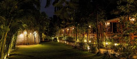 Une vue nocturne d’un hôtel, avec un chemin bien éclairé, entouré d’une végétation luxuriante et de palmiers.