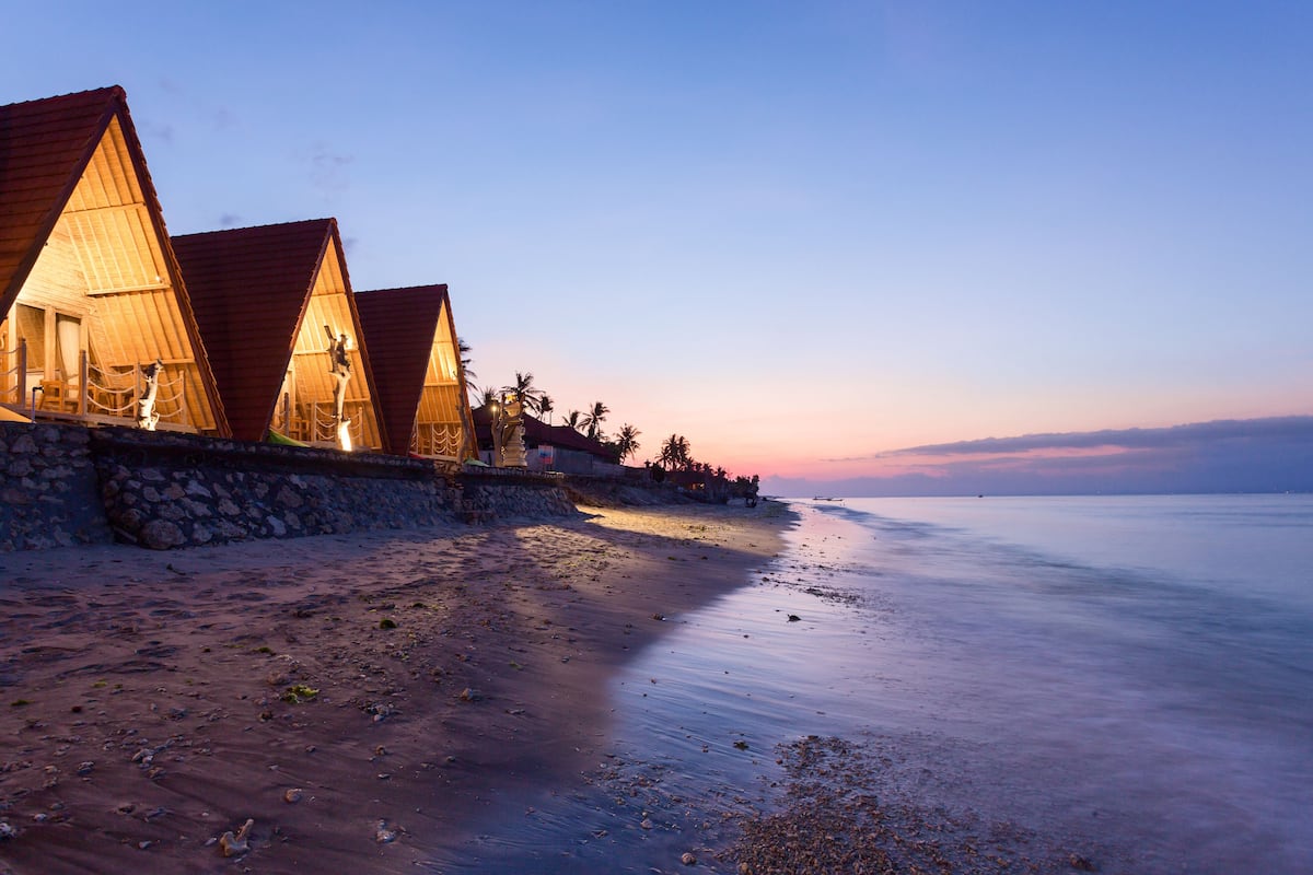 A beachfront property with wooden triangular-roofed cabins, a stone wall, and a calm sea.