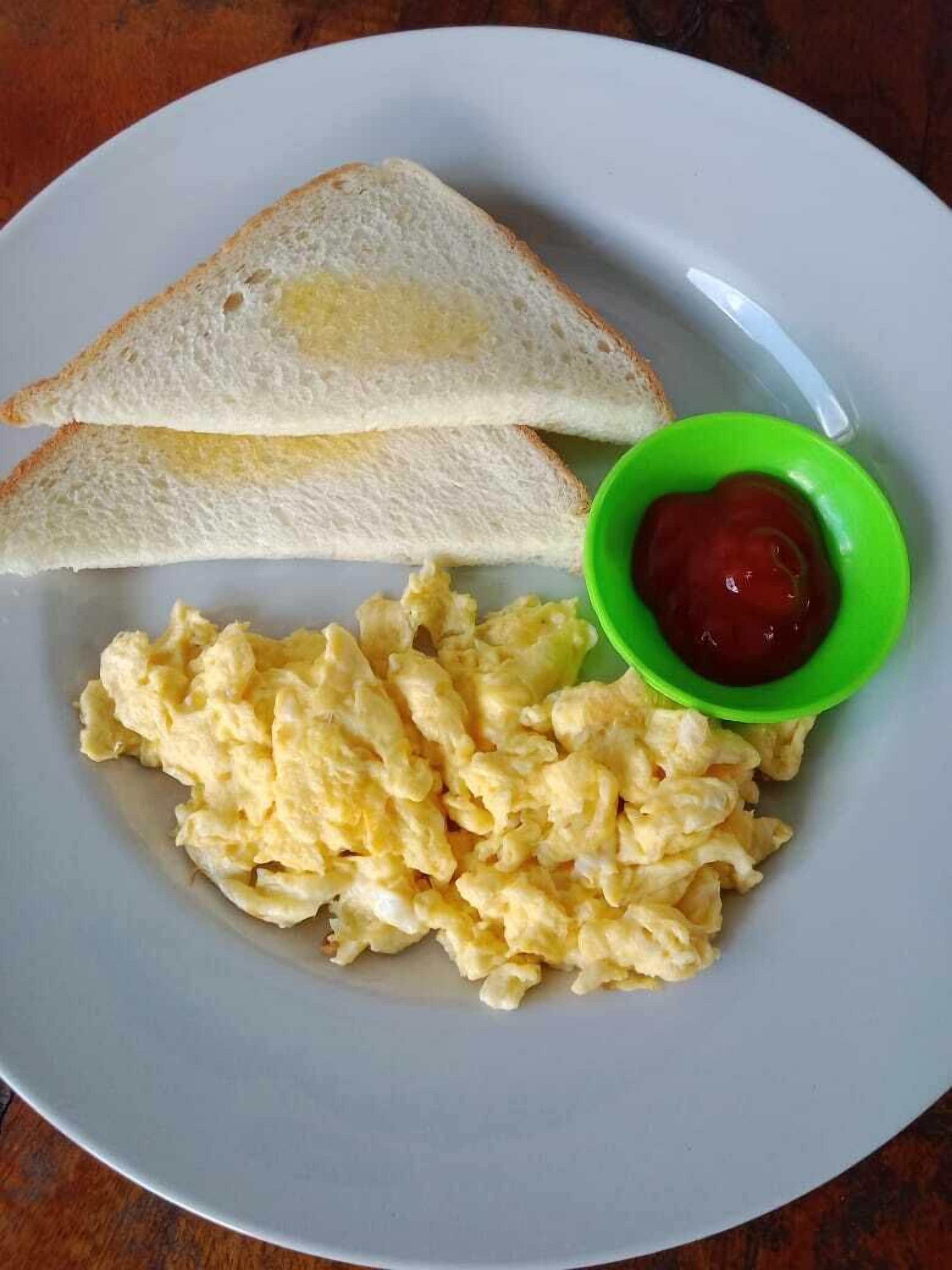 A plate with scrambled eggs, two slices of toast, and a small bowl of ketchup.