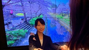 Une personne est assise à une table, entourée de documents, devant un grand écran qui affiche un paysage pittoresque.