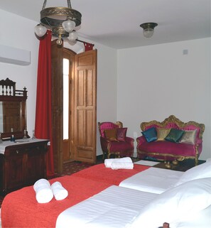 A hotel room with a wooden door, a red curtain, a chandelier, two ornate red and gold armchairs, a wooden dresser, and a bed with white linens.