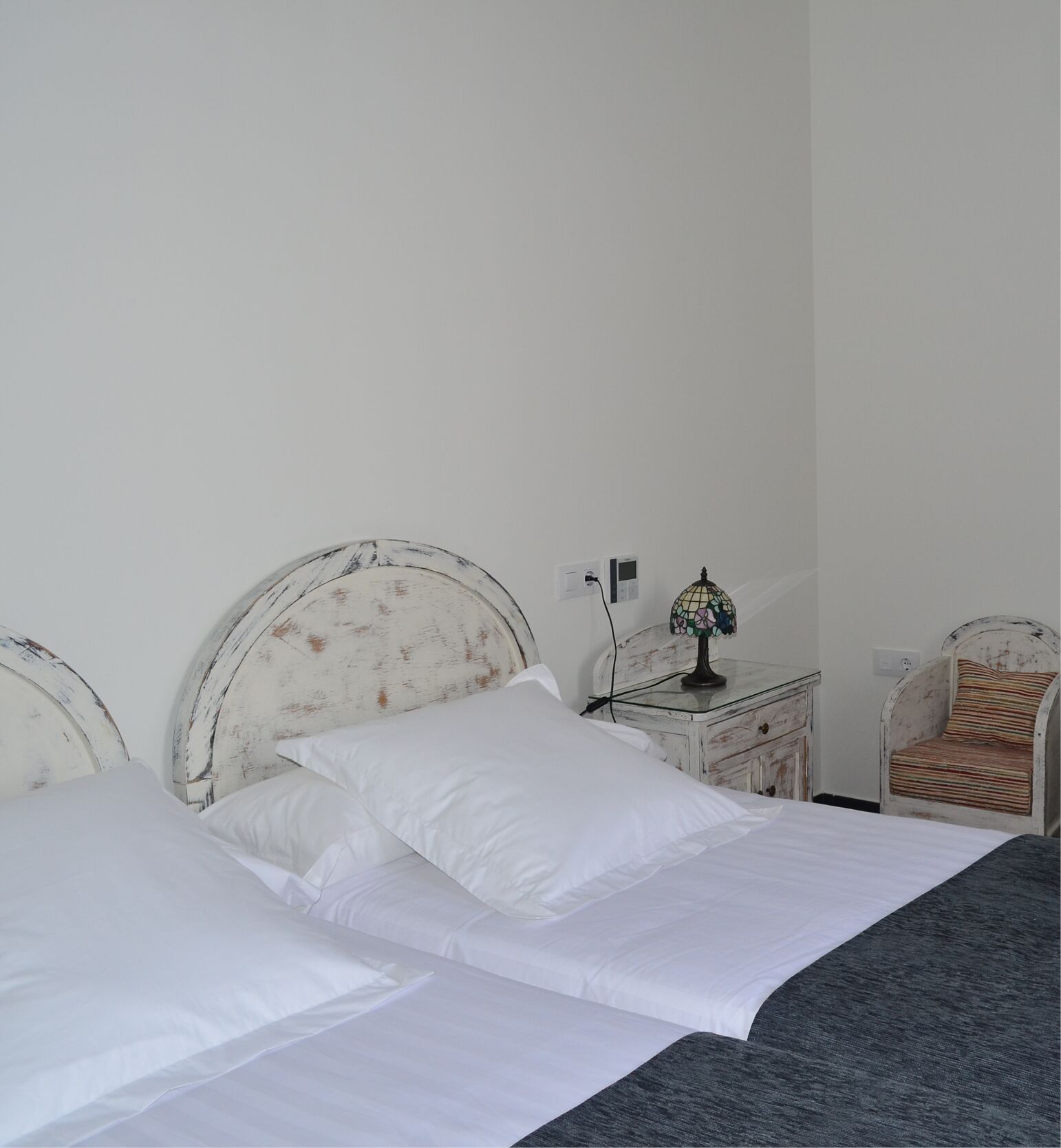 A bed with white bedding and a grey blanket, a small bedside table with a lamp, and a chair with a striped cushion.