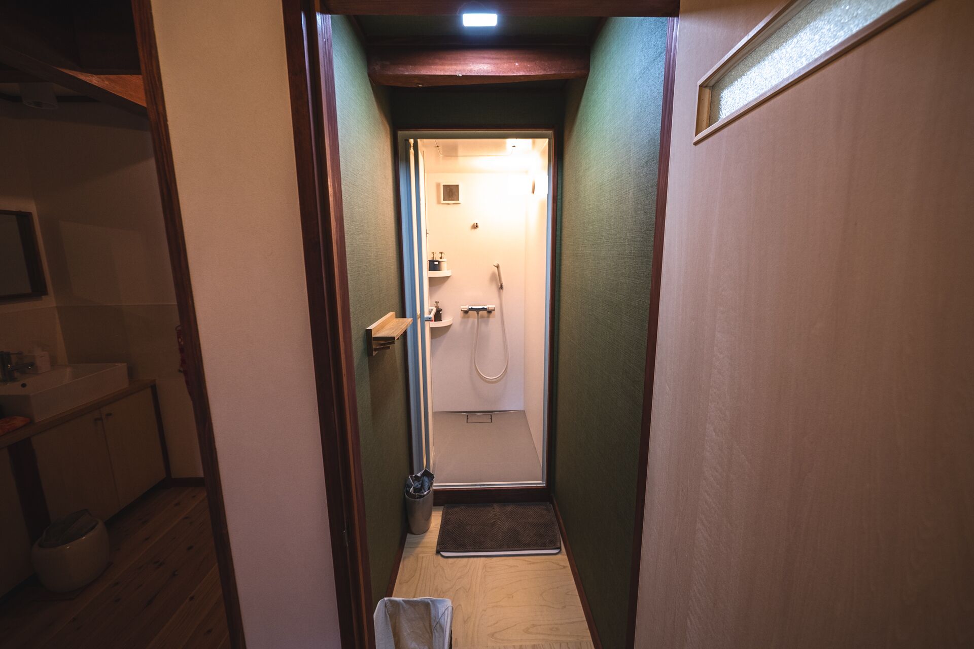 A narrow hallway leading to a bathroom with a shower, a small shelf with toiletries, and a wooden floor.