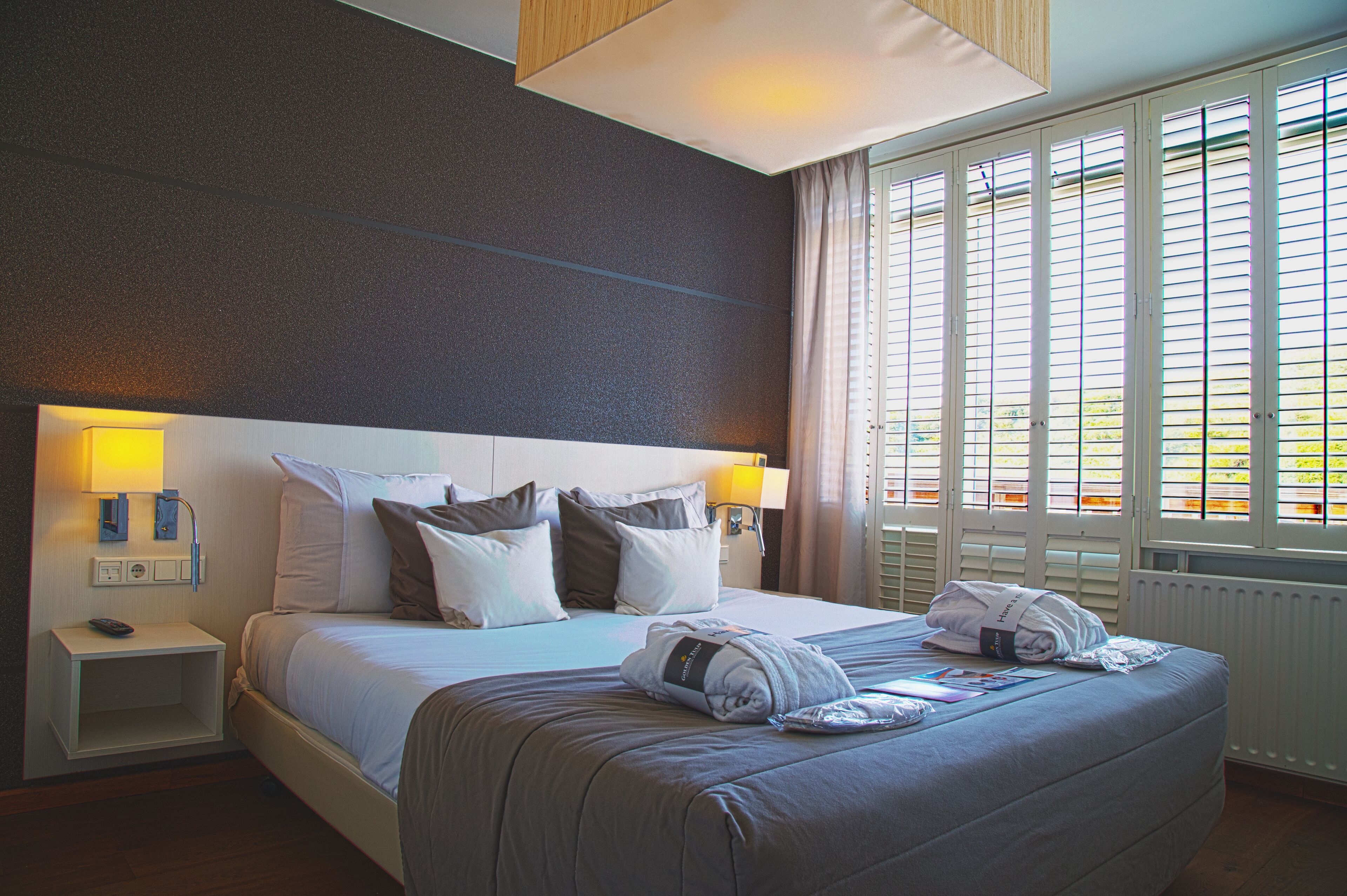 A hotel room with a large bed, two bedside lamps, a nightstand, and a window with blinds.