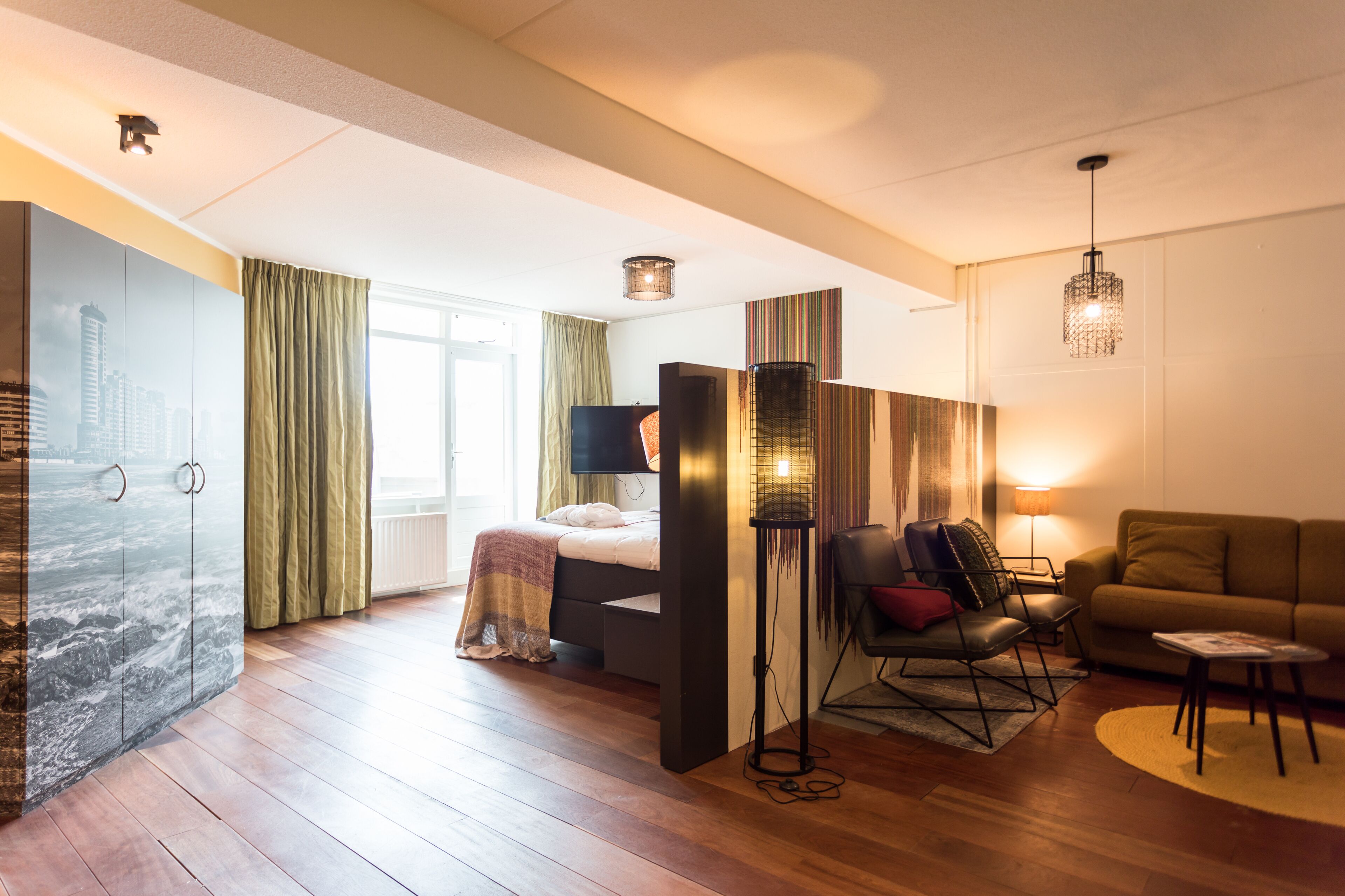 A modern hotel room with a large bed, a seating area with a sofa and chairs, a floor lamp, and a large window with curtains.