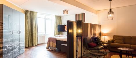Une chambre d’hôtel moderne dotée d’un grand lit, d’un coin salon avec un canapé et des fauteuils, d’une lampe sur pied et d’une grande fenêtre avec des rideaux.