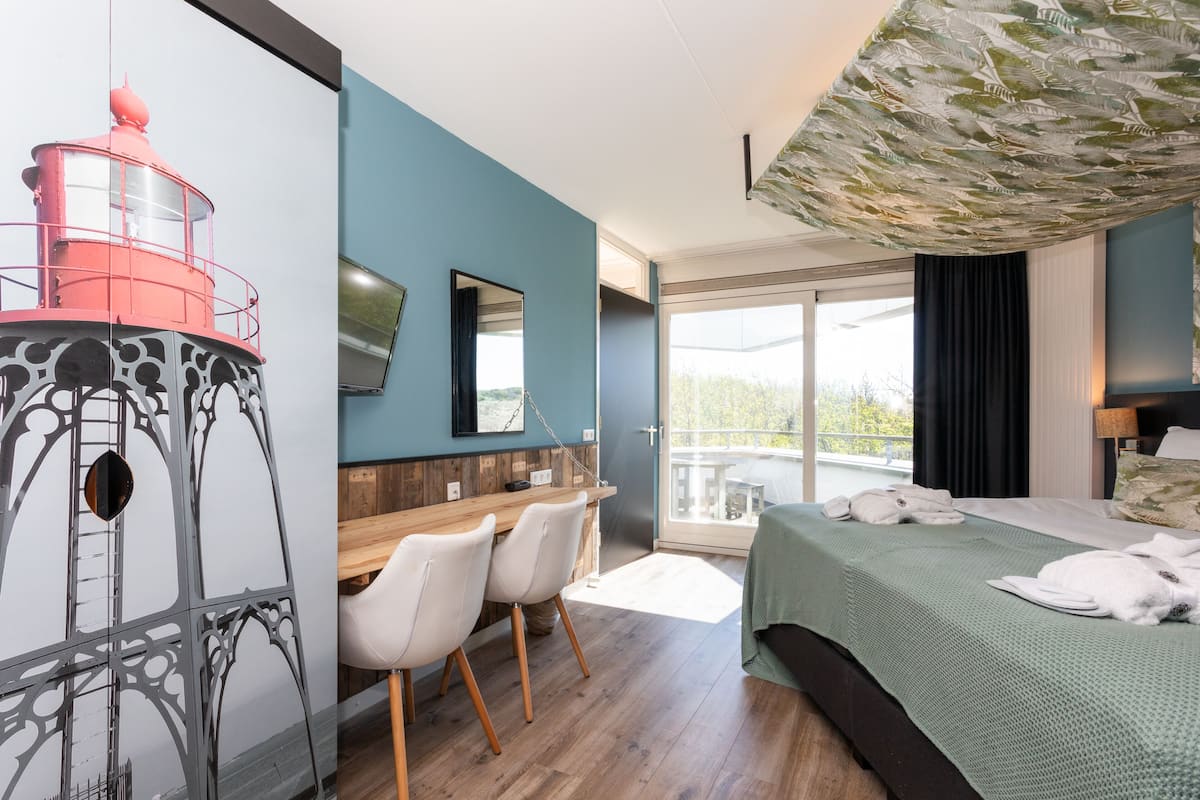A modern hotel room with a large bed, a dining table with chairs, a balcony with a view, and a unique wall decoration.