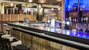 A bar area with stools, a bar counter, and a shelf with bottles. - Strandhotel Westduin (Koudekerke)