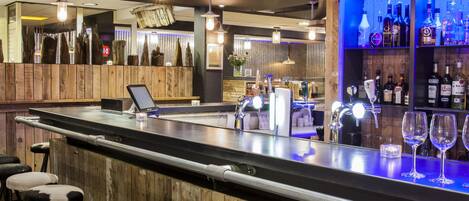 Una zona de bar con taburetes, mostrador y estantería con botellas.