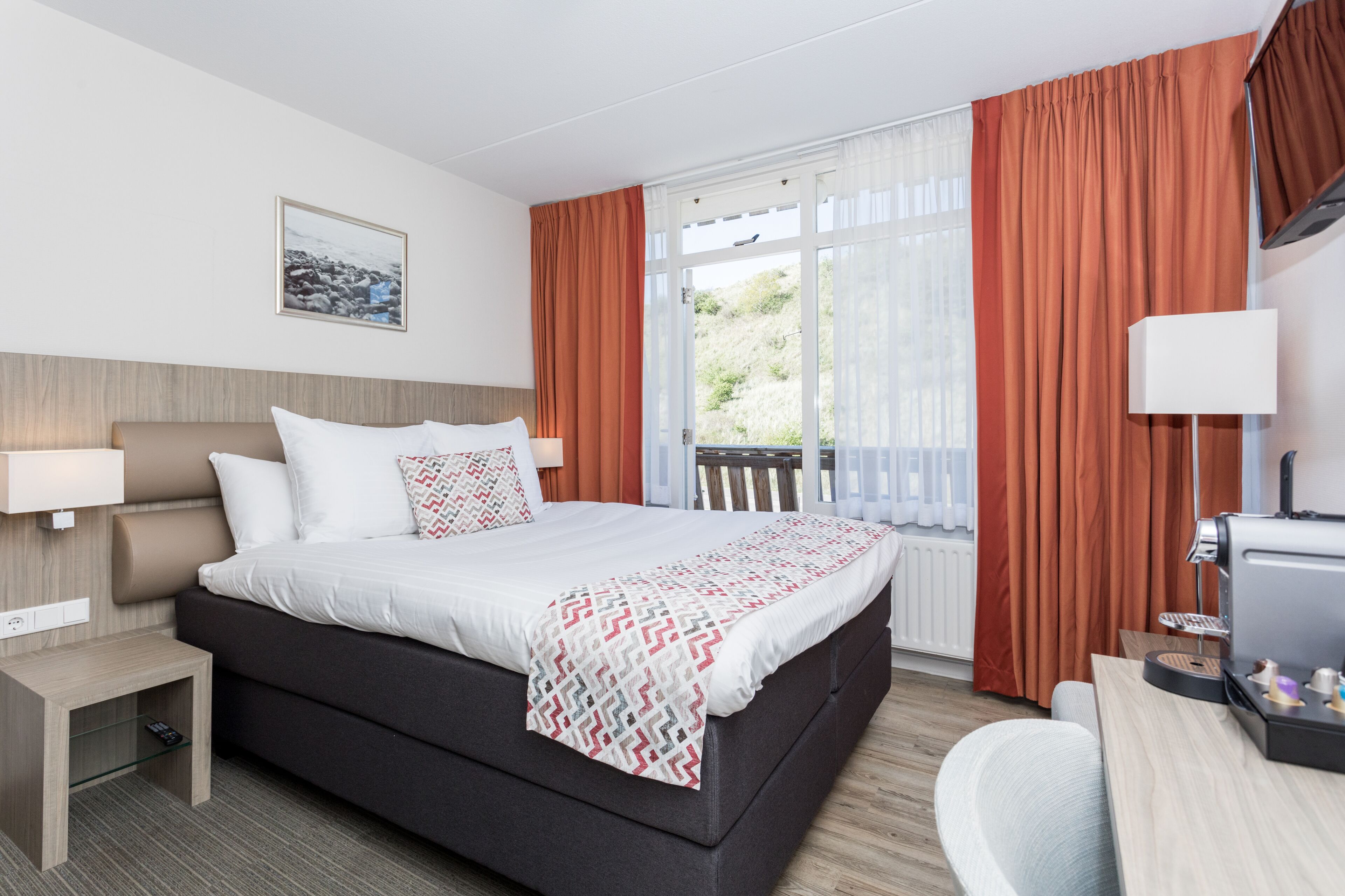 A hotel room with a bed, bedside table, lamp, and a view of the outdoors.