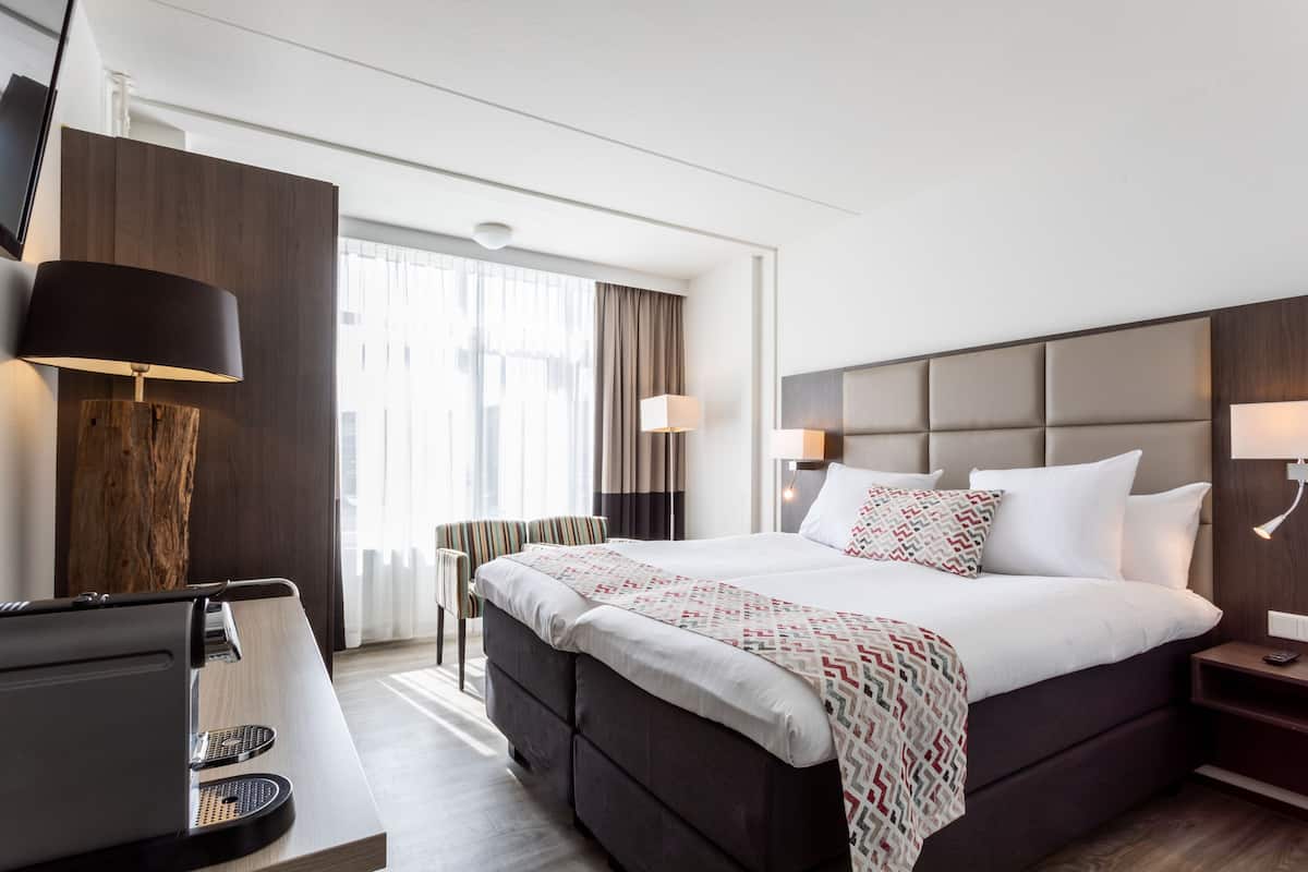 A modern hotel room with a large bed, a desk with a coffee machine, and a TV on the wall.