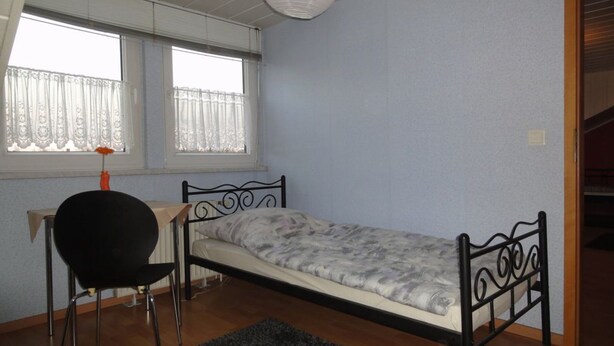 Une petite chambre avec un lit simple, un cadre en métal, une chaise et un radiateur.