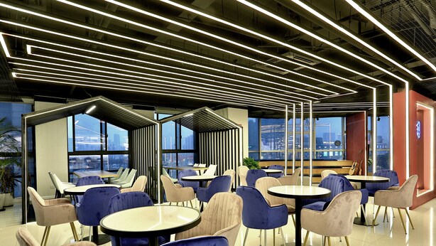 A modern cafe with round tables and chairs, large windows offering city views, and a ceiling with linear light fixtures. - Hangzhou Infinity Youth Hostel (Hangzhou)