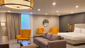 Une chambre d’hôtel comprenant un lit, deux fauteuils, un canapé, un miroir rond, une lampe sur pied et un bureau.