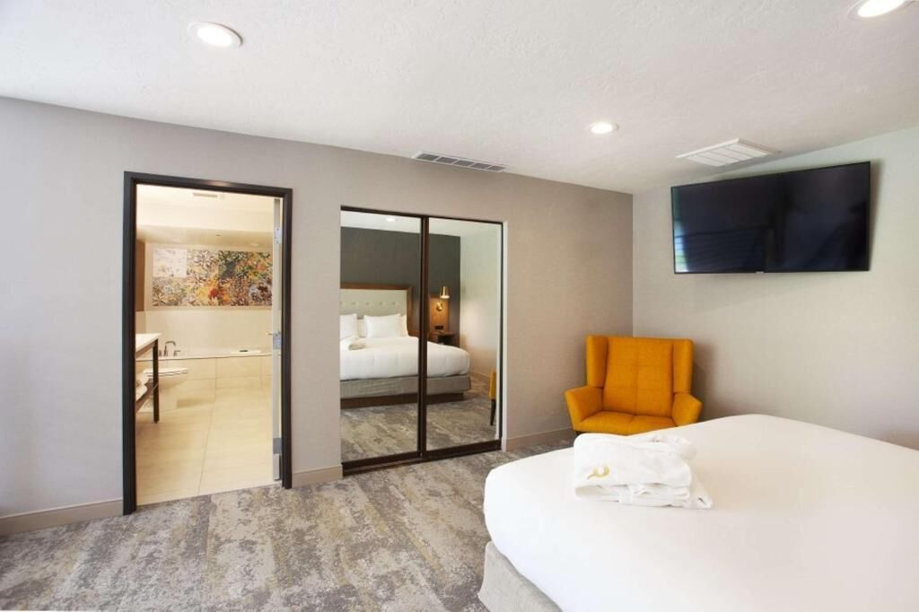 A hotel room with a bed, a TV mounted on the wall, and a bathroom visible through a glass door.