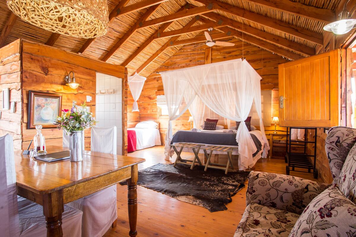 Una cabaña rústica de madera con una cama bajo una dosel, una mesa de comedor con sillas, y una zona de estar acogedora con tapicería estampada.