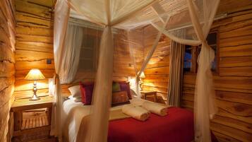 A wooden cabin room with a bed, a nightstand, a lamp, and a window with curtains.