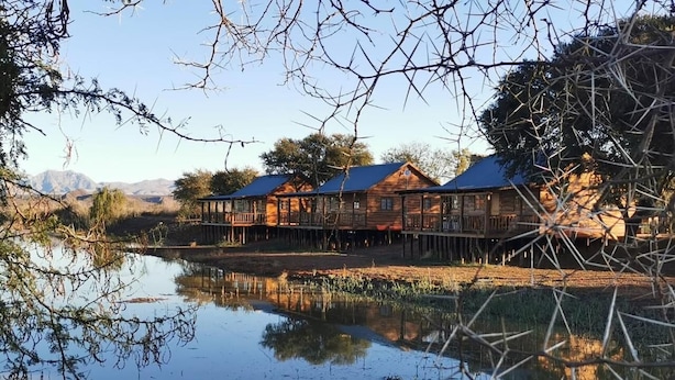 A row of wooden cabins with a reflective water body in the foreground. - Soetdoring Selfsorg (Oudtshoorn)