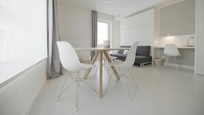 A modern, minimalist living room with a dining table, white chairs, a grey sofa, and a small desk with a lamp. - Florence en Ville (Brussels)