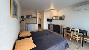 Habitación de hotel pequeña con cama, mesitas de noche, escritorio, silla, kitchenette y televisor.