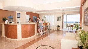 A hotel lobby with a reception desk, a seating area with a view, and a balcony with outdoor seating.