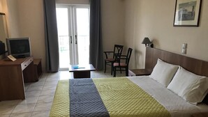 A hotel room with a bed, a desk with a television, a chair, and a window with curtains. - Ariadne Apartments (Zakynthos)