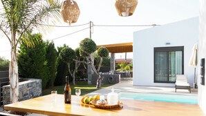 Uma área de refeições ao ar livre com uma mesa de madeira, cadeiras e piscina ao fundo.