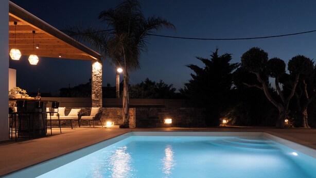 Piscina com parede de pedra e área de estar coberta, com cadeiras e uma mesa.
