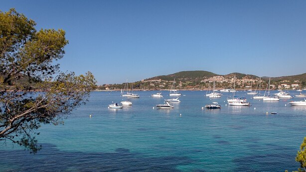 A coastal marina with various boats anchored in clear blue waters, surrounded by rocky terrain and greenery.