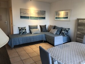 A cozy room with a sectional sofa, a patterned table, and artwork depicting a beach scene. - Studio near beach, shops and station with balcony, parking and wifi (Saint-Raphaël)