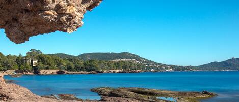 A coastal landscape with rocky terrain, clear blue water, and distant hills.