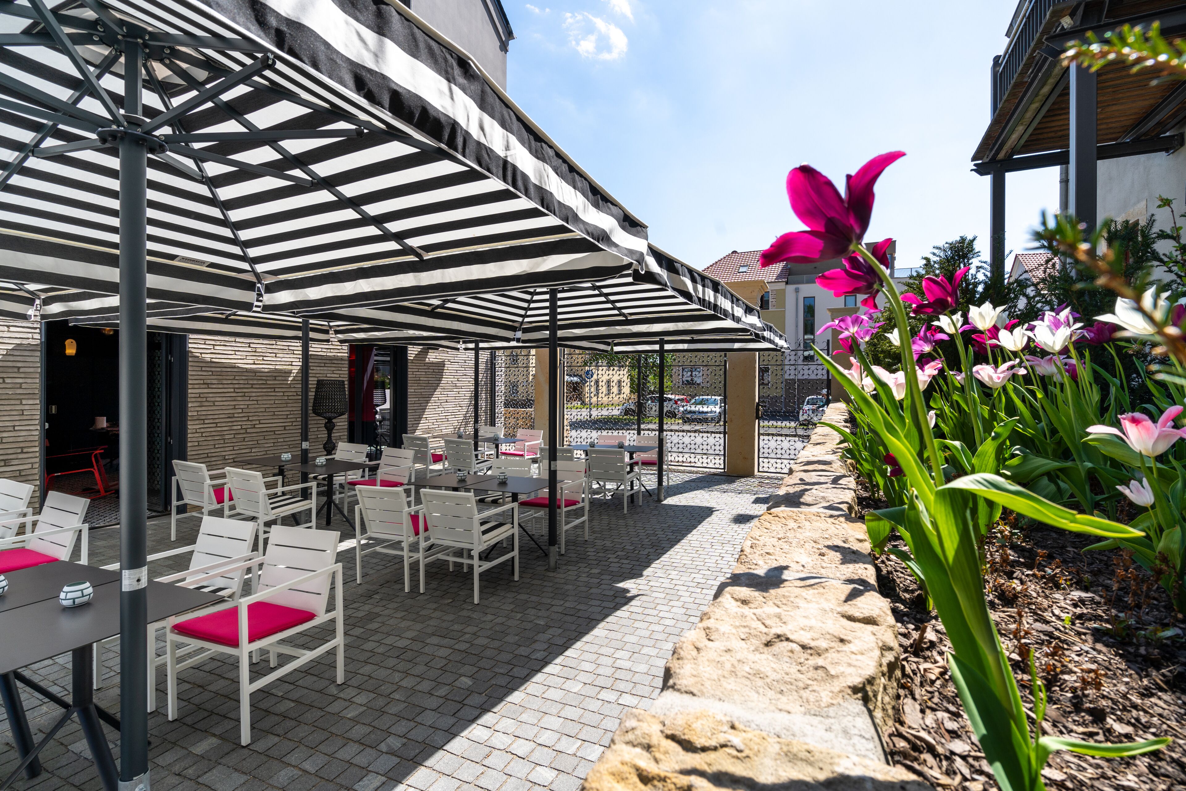 Un patio al aire libre con sillas y mesas blancas, cubierto por sombrillas a rayas, rodeado de un muro de piedra y plantas con flores.
