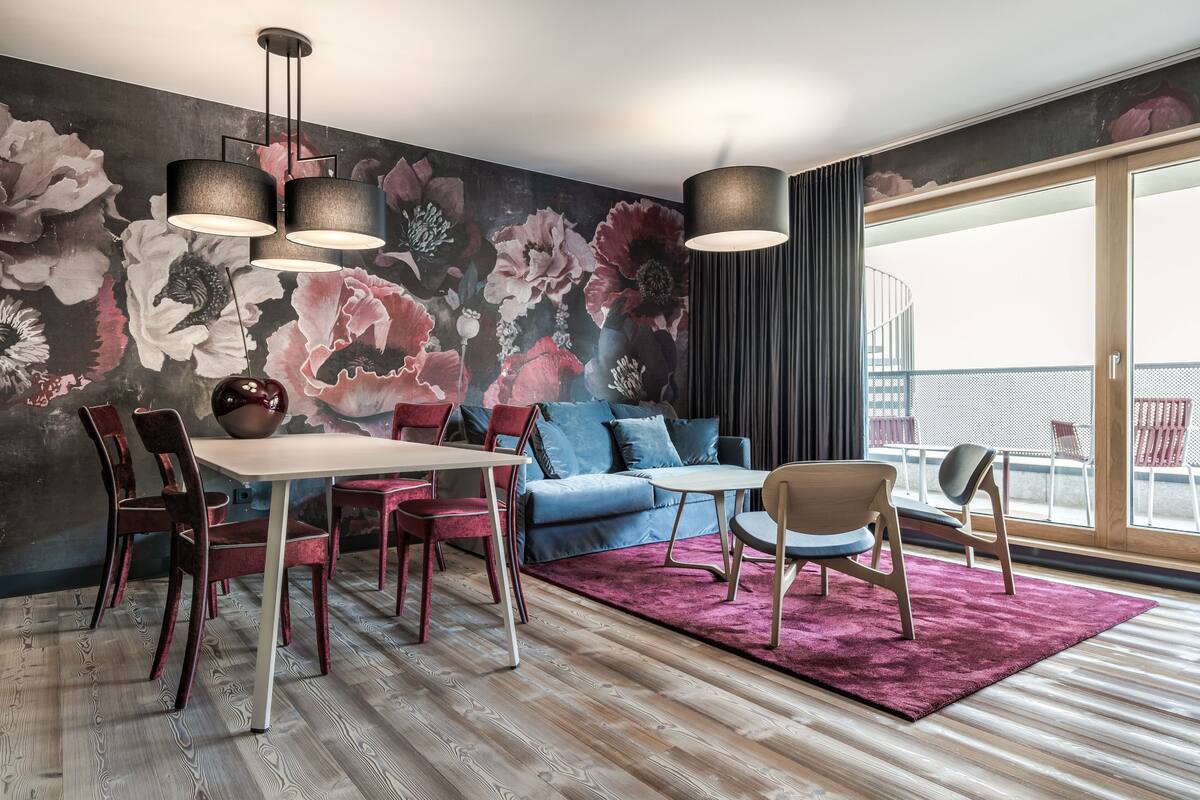 Un salon moderne avec un coin repas, agrémenté d’une grande fresque florale, d’une table à manger en bois avec des chaises, d’un canapé bleu et d’une grande fenêtre offrant une vue sur la ville.