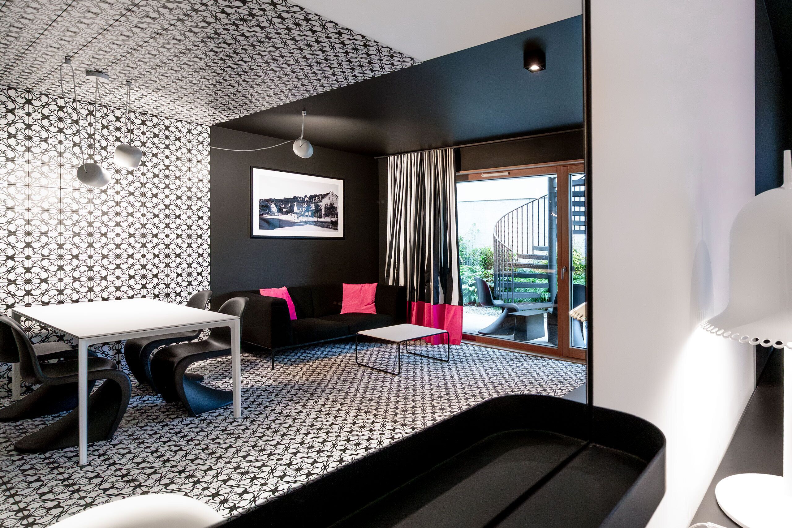 Una habitación moderna con suelo estampado, un sofá negro con cojines rosados, una mesa blanca y un ventanal con cortinas.