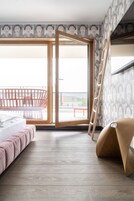 A modern hotel room with a large sliding glass door leading to a balcony, a bed with a pink headboard, and a wooden ladder leaning against the wall.