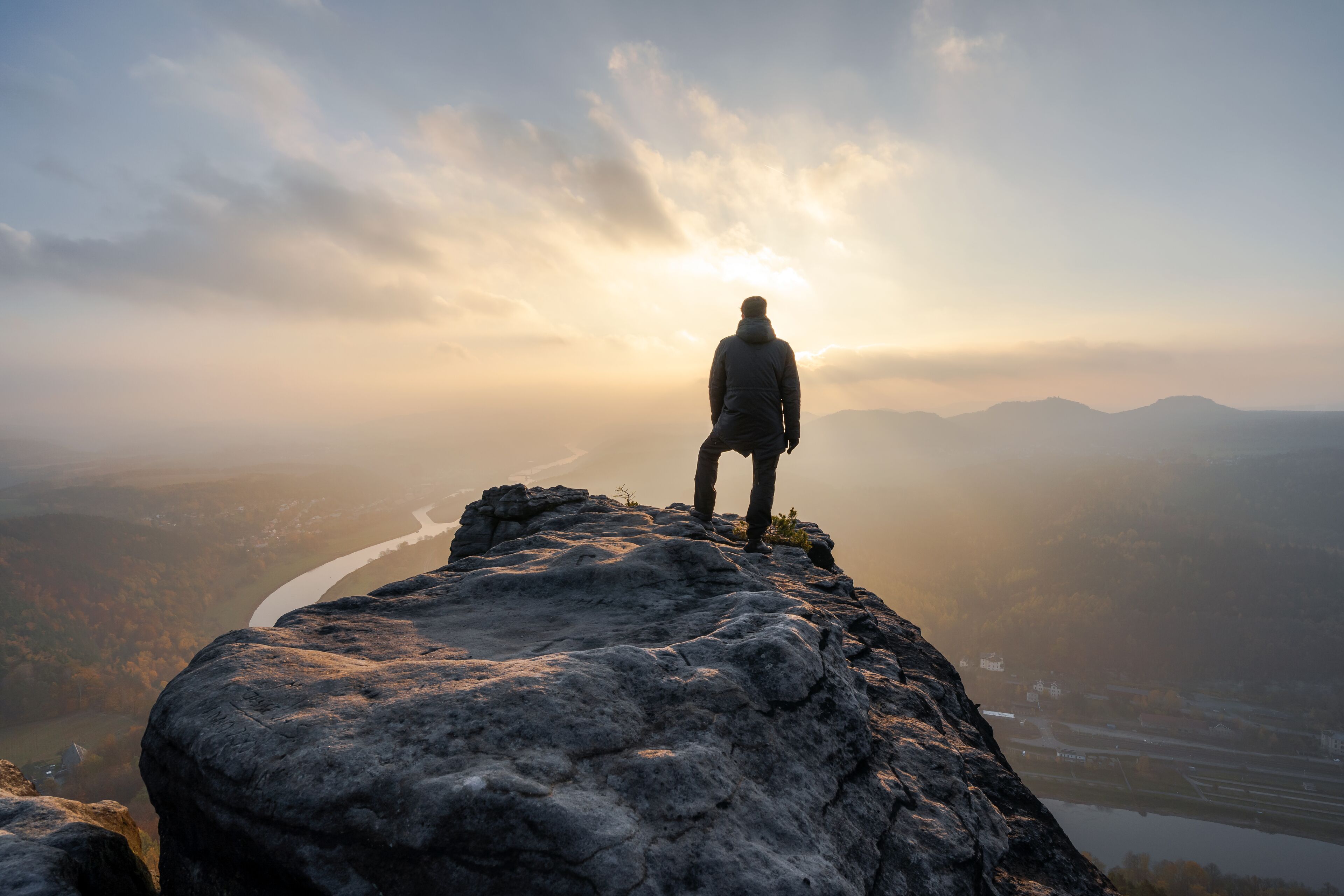 A person standing on a rocky outcrop overlooking a valley with a river and distant mountains.