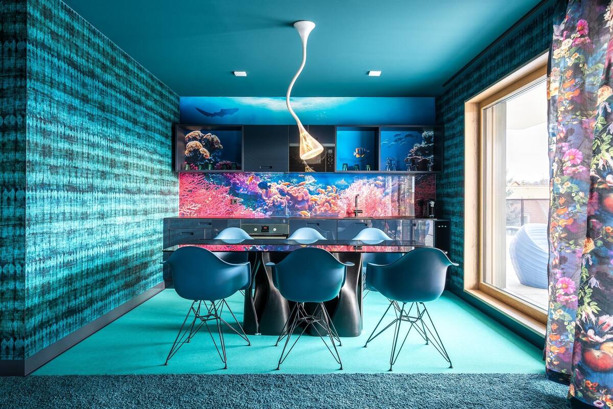 Une cuisine moderne dotée d’un grand îlot central, de chaises bleues et d’une fresque murale aux couleurs vives, dans les tons corail.
