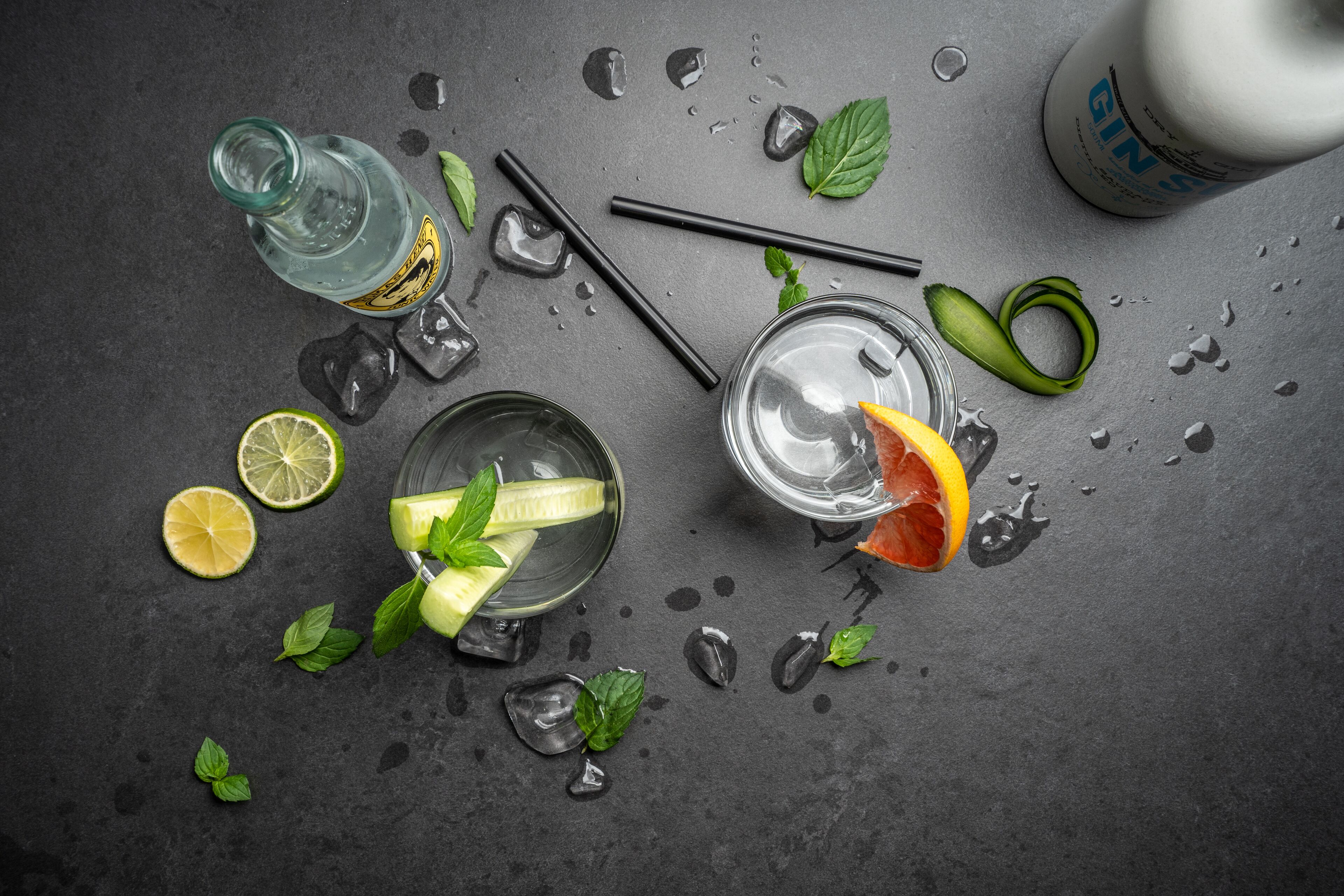Ein Glas Wasser mit Orangenscheibe und Minzblättern, ein Glas mit Limettenscheibe, eine Gin-Flasche und Eiswürfel auf einer dunklen Oberfläche.