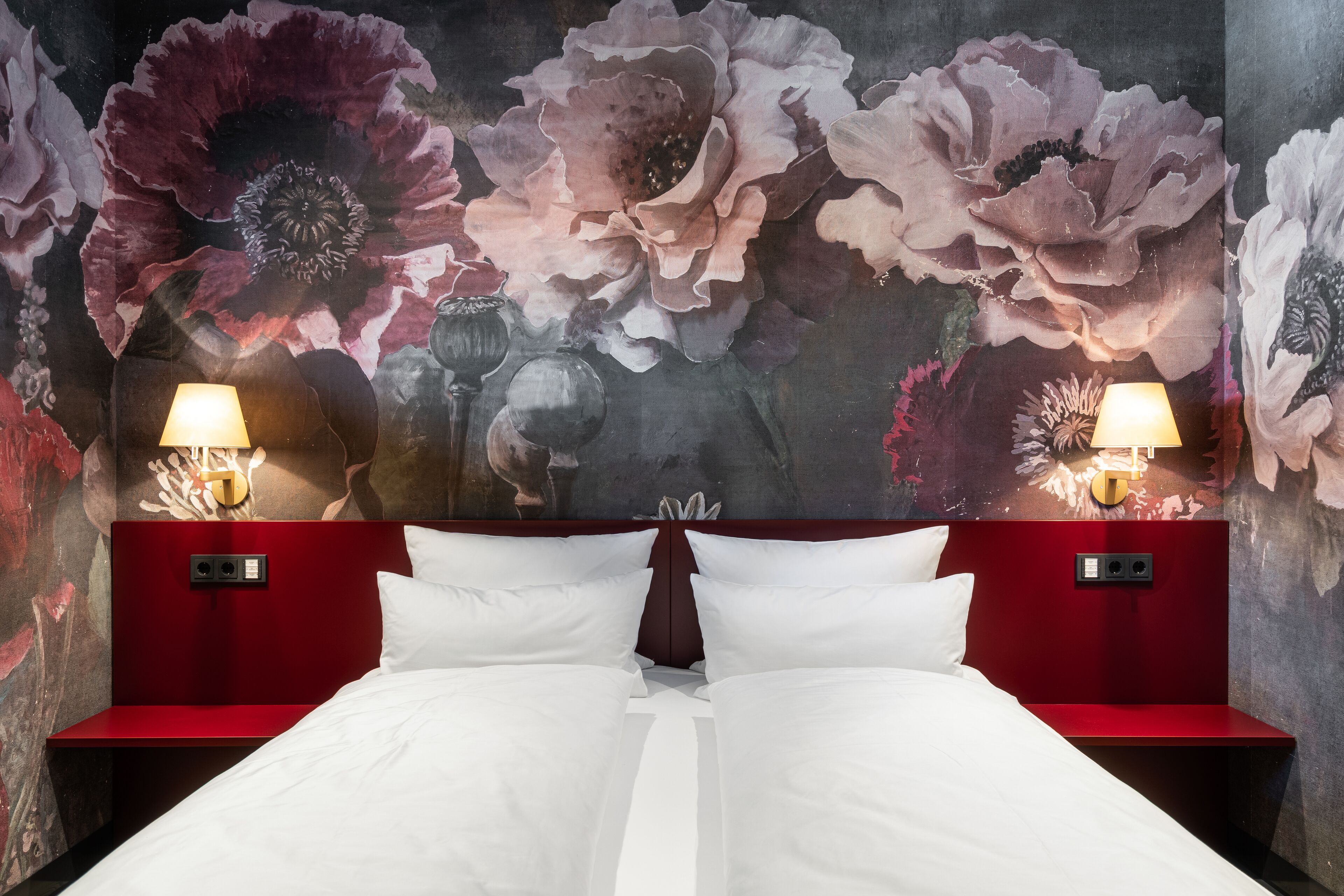 A double bed with white linens, two bedside tables with lamps, and a floral mural on the wall.