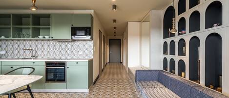 A narrow hallway with patterned flooring, a kitchen area with green cabinets and a microwave, and a seating area with a patterned bench.