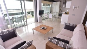 Ein modernes Wohnzimmer mit einer Couch, einem Couchtisch und einem Fernseher. Es gibt einen Balkon mit Blick ins Freie.