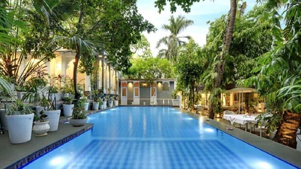 An indoor swimming pool surrounded by lush greenery and a building with arched windows. - Hotel Raj Palace (Udaipur)