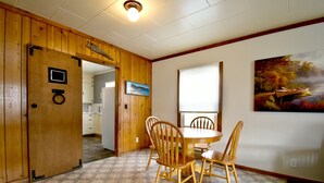 A dining area with wooden chairs and a table, a kitchen in the background, and a painting on the wall. - Coffee Klatch Cottage - Yellow (Potlatch)