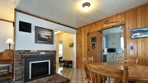 A dining area with a brick fireplace, wooden dining table, and chairs. A kitchen is visible in the background. - Coffee Klatch Cottage - Yellow (Potlatch)