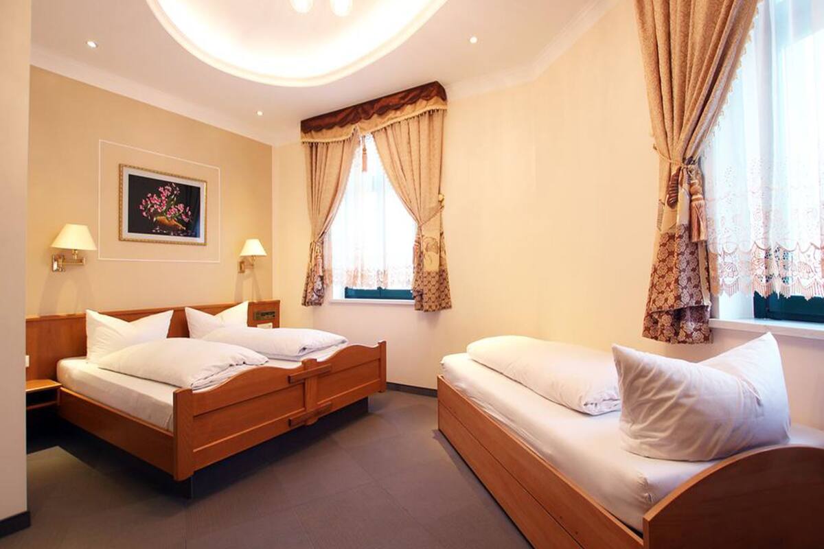 A hotel room with a double bed, a single bed, two bedside tables, a chandelier, a framed picture on the wall, and a window with curtains.